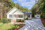 A charming single-story house at 4080 Brooksdale Drive in Franklinton boasts a light-colored exterior, embraced by lush trees. It features a welcoming front porch, neatly trimmed bushes, and a spacious driveway. A small shed peeks from the background under the partly cloudy sky.
