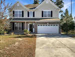 This two-story house at 2205 Hyde Court in Creedmoor boasts light beige siding and brick accents. It features a white two-car garage, black shutters, and a manicured front lawn with small shrubs. The concrete driveway leads up to the home, set against a backdrop of clear blue skies and trees.