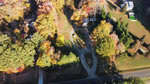 Aerial view of the rural landscape near 2037 Mountain Creek Road in Oxford, capturing autumn's vibrant palette. A tree-lined road meanders past scattered houses and a green pool, while the foliage in hues of orange, yellow, and red casts long shadows over the grassy terrain.