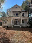 A large, old, abandoned house with peeling white paint stands surrounded by trees at 217 College Street. It has a two-story porch with columns, leaves scattered on the ground, and a clear, sunny Oxford sky above.