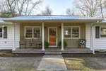 A cozy white house at 2611 Tar River Road in Creedmoor boasts a front porch with two rocking chairs and a small table on the left, and a bench draped with a blanket on the right. Adorned with plants and a door wreath, its charm is enhanced by the metal roof and tree-lined backdrop.