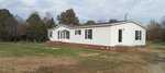 Located at 4142 Deer Run Drive in Oxford, this cozy single-story white mobile home rests on a brick foundation amidst a grassy lot. Featuring multiple windows and a charming front porch, it is beautifully framed by trees under a partly cloudy sky.