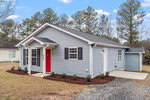 Nestled on 503 Cotton Avenue in Creedmoor, this charming single-story gray house boasts a gabled roof with a bright red front door and crisp white trim. The modest yard features a few small shrubs, while tall trees stand proudly against the clear blue sky backdrop.