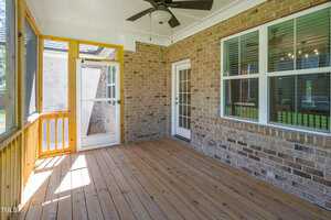 Bright sunroom with wooden flooring and brick walls, featuring a ceiling fan. Large windows and a screen door provide ample natural light and outdoor views.