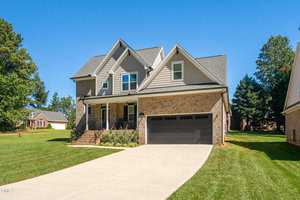 A charming two-story house at 407 Eagle Court in Henderson, showcasing a brick facade and gray siding. It features a large gable roof, a covered front porch, and a black garage door. The neatly trimmed lawn is set against a backdrop of trees and a clear blue sky.