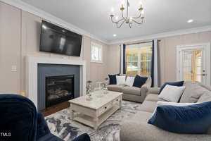 Living room with a modern design, featuring a mounted TV above a fireplace. The space includes a marble-top coffee table, gray and navy seating, and a patterned area rug. Natural light streams in through windows with curtains. A chandelier hangs from the ceiling.