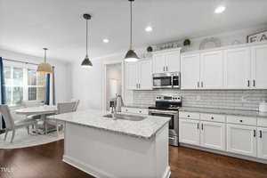 Modern kitchen with a granite island and stainless steel appliances, including a stove and microwave. White cabinets with black handles line the walls. A dining area with a round table and chairs is visible near large windows with blue curtains.
