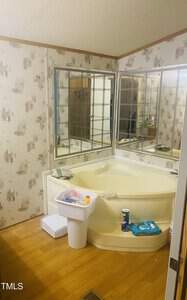 Bathroom with a corner bathtub, surrounded by large mirrors. The walls have a rustic pattern, and the floor is wooden. Toiletries and cleaning supplies are on the tub ledge and nearby trash bin.