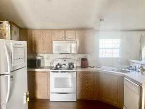 A kitchen with wooden cabinets, a white refrigerator, stove, microwave, and dishwasher. Cooking pots are on the stove, and a window is above the sink, letting in natural light. The fridge has papers and notes attached to it.