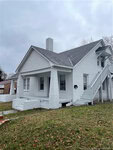 The two-story, white brick house at 410 E Montgomery Street features a covered porch and an external staircase to the upper level. The residence, located in Henderson, has a gabled roof and chimney, all surrounded by a grassy yard with scattered fallen leaves under an overcast sky.
