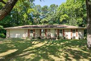 Nestled at 101 Jeffress Drive in Henderson, this single-story brick house features a light brown roof and is surrounded by tall trees. The large garage on the left complements several front windows, while a well-maintained lawn and a few plants adorn the welcoming entrance.