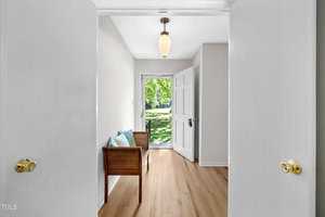 A bright hallway with light wood flooring and white walls leading to a doorway with a view of a green lawn outside. A wicker bench with blue cushions is on the left side, and a hanging light fixture is above.
