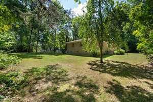 A small house with light-colored siding stands in a wooded area, surrounded by trees. The grassy lawn is sunlit, creating a peaceful and secluded atmosphere. Shadows of trees are cast on the lawn, illustrating a bright, sunny day.
