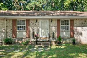 A single-story brick house with a brown shingle roof, white-framed windows, and brown shutters. A small porch with metal railings leads to the front door, surrounded by greenery and trees in the background on a sunny day.