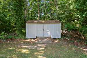A small, light gray shed with a sloped roof stands in a wooded area. Surrounded by dense trees and foliage, the shed has a front door secured with a metal latch. Sunlight filters through the trees, casting dappled shadows on the ground.