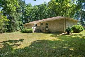Single-story brick house with a sloped roof, surrounded by trees and a grassy lawn. The backyard features shrubs and a small deck with steps leading to a door.
