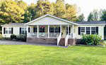 Located at 3128 Henry Wilson Road in Oxford, this single-story house boasts light yellow siding and a sturdy brick foundation. The covered porch is adorned with hanging plants, while the well-maintained yard features a lush green lawn and scenic trees as its backdrop.