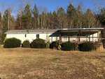 Nestled on 8178 Raymond Royster Road in Roxboro, this single-story house features white siding and a charming wraparound porch, beautifully set against a backdrop of tall trees. The grassy front yard is dotted with bushes, all under a clear blue sky.