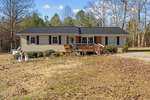 Located at 8019 Harts Cross Road, this charming one-story house in Oxford features a dark gray roof and light siding, nestled among trees. A wooden deck with railings graces the front, adorned with decorative snowmen and poinsettias. The yard's dry grass is dotted with scattered leaves.
