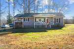 A small gray house at 3202 Little Mountain Creek Road, Oxford, boasts a front porch decorated for the holidays with a wreath and red ribbon on the door. The brown lawn, framed by trees, sits under a clear sunny sky, while a driveway and additional structure are visible to the left.