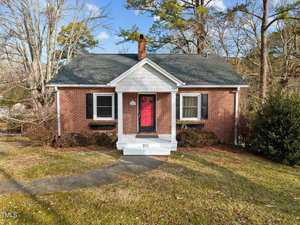 Nestled on 211 Park Avenue, this small, charming brick house with a gabled roof and a red front door captures the essence of Creedmoor. Surrounded by trees, it features white trim, black shutters, and a concrete path leading to the entrance. The grassy lawn is complemented by shrubs near the facade.
