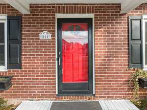 A brick house exterior with a bright red front door framed by black shutters on either side. The house number, 211, is displayed above the door. A small planter with greenery accents the entrance.