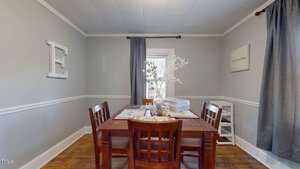 A dining room with a wooden table and four chairs on a wooden floor. The room has gray walls with white trim and a large letter "E" on one wall. A window with dark curtains brightens the space, and a white vase with branches decorates the table.