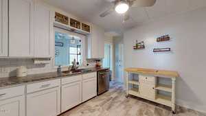 A modern kitchen with white cabinets, granite countertops, and a stainless steel dishwasher. A small kitchen island with shelves and a cutting board is on the right. Overhead, a ceiling fan with a light illuminates the room.
