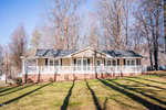 The single-story tan house at 2665 Gela Road in Oxford features a black roof and white trim, along with a long front porch. Surrounded by leafless trees under a clear blue sky, it boasts a well-kept grassy lawn in the foreground.