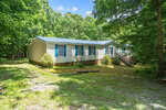 Nestled on 3196 Loblolly Lane in Wake Forest, a beige mobile home with blue window shutters sits peacefully in a wooded area. It features a metal roof, brick underpinning, and a small front staircase. Tall trees encircle the home, while grass and gravel mix under a clear blue sky.