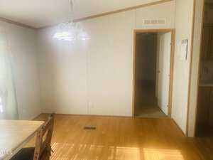 A sunlit room with wooden flooring, featuring a hanging light fixture above a dining area. A wooden chair is partially visible on the left. The walls are light-colored with a doorway leading to a darker room on the right.