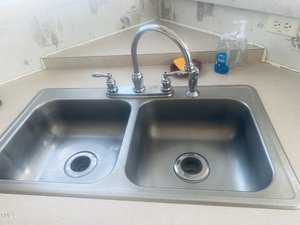 A stainless steel double kitchen sink with a chrome faucet. There are cleaning supplies, including a hand soap dispenser and a sponge, on the countertop. The backsplash is decorated with light-colored wallpaper.
