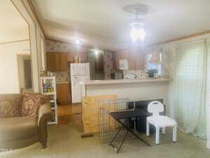A cozy living room with beige carpeting, featuring a brown sofa on the left. An open kitchen with wooden cabinets and white appliances is in the background. A small table and a child's chair are positioned near a curtained window on the right.