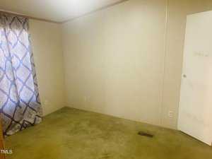 An empty room with beige walls and a closed white door. A single window is covered by a curtain with a blue and white pattern. The floor is carpeted in a light green color. The room appears bare and undecorated.