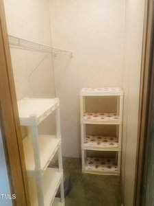 A narrow storage room with beige walls features white plastic shelving units on both sides. The shelves have floral-patterned liners. A metal wire rack is mounted on the left wall. The floor appears to be concrete.