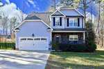 The charming two-story house at 2163 Emerald Lane in Franklinton boasts gray siding and stone accents. It features a double garage door, windows with shutters, and a well-maintained lawn. Trees stand gracefully in the background under a blue sky with scattered clouds.