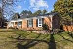 A single-story brick house on 500 Sunset Avenue in Oxford features white trim and a covered front porch with rocking chairs. The yard is grassy, with a few trees casting long shadows. Pine trees stand in the background under a blue sky with scattered clouds.
