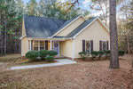 A beige house at 3714 Aztec Lane, Franklinton, with a dark roof is surrounded by tall pine trees. It features dark shutters, a front door with a wreath, and two windows adorned with wreaths. A concrete path leads to the entrance, and the ground is covered with pine needles.