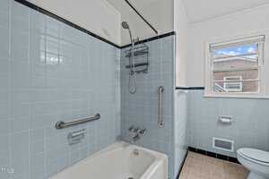A bathroom featuring light blue tiled walls, a white bathtub with a showerhead, and a window with a view of a building outside. The bathtub includes a grab bar and built-in soap holder. The floor is tiled in beige.