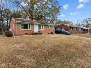 A brick house with a white door and green window shutters sits on a grassy lawn. A black tarp covers an object in the driveway. A similar house is visible nearby. Trees and a blue sky with clouds are in the background.