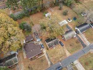 Aerial view of a suburban neighborhood with brick houses surrounded by autumn trees. Cars are parked in driveways, and a trampoline is visible in a backyard. The streets form a grid pattern, and the grass is mostly brown.