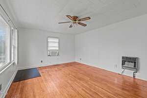 A spacious, empty room with white walls and a wooden floor. There's a ceiling fan, a window air conditioning unit, and a wall-mounted heater. Large windows on the left allow natural light to fill the room.