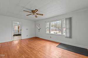 A bright, empty living room with wooden floors and a ceiling fan. Large windows on the right let in natural light. An open doorway leads to another room, hinting at a kitchen area with visible windows.