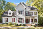 The charming two-story white house at 567 Timberknoll Lane, Wake Forest, boasts a gabled roof and red shutters. Surrounded by trees, it features a front porch with columns and a brick foundation, with manicured bushes lining the front. The clear blue sky suggests a sunny day.