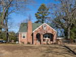 Nestled at 3257 Tump Wilkins Road in Stem, a brick house with a green roof, arched windows, and a central chimney stands amid a yard with bare trees. The sky is clear and blue, with trees and power lines silhouetted in the background.