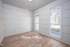 A bright, empty home office with light wooden floors and white paneled walls. Large windows on the right let in natural light, and there's a ceiling light fixture. Text overlay reads "Home Office" and "2.7.2024.