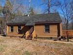 A small log cabin nestled at 4068 Culbreth Road features a dark shingled roof, two front windows, and a central door. It has a wooden porch with steps leading up to the entrance. Surrounded by bare trees and a newly planted garden bed, the cabin also boasts a deck visible on one side.