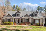 Nestled at 1078 Anjanette Lane, this single-story gray house with dormer windows and a stone facade is surrounded by a mix of evergreen and deciduous trees. The front yard in Creedmoor is grassy, with small shrubs near the entrance, under a clear sky dotted with a few clouds.
