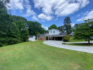 A large house with white siding and multiple levels is surrounded by green lawns and tall trees under a partly cloudy sky. A paved driveway leads to a carport, and a small white shed is visible in the yard.