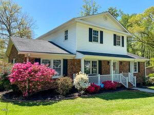 A two-story house with a brick and white siding exterior. The front porch has a white railing, and the yard features colorful blooming bushes, including pink and red flowers. Tall trees surround the property under a clear blue sky.